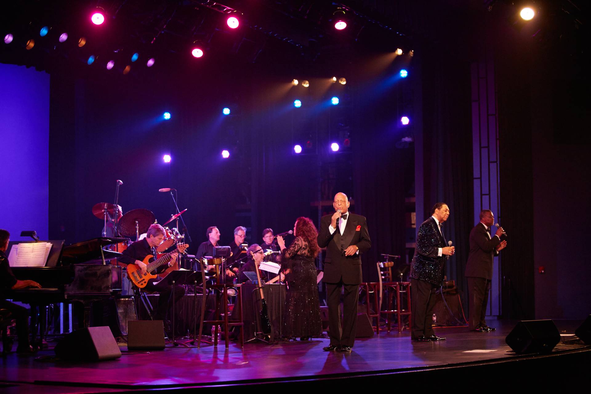A vocalist and full band performing under stage lights on the Highlander Auditorium stage